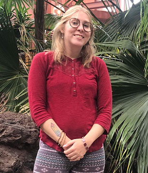 Portrait: Gillian Corsiatto glasses and a red shirt standing in front of a palm tree.